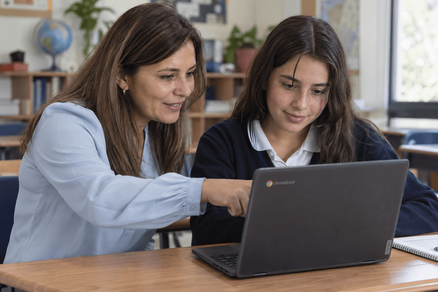 Escena creada con IA: una docente guía a un estudiante usando un Chromebook.